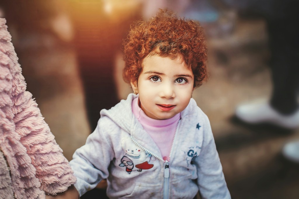 Curly-haired child in a light hoodie looks at the camera while holding an adult's hand in a softly lit setting.