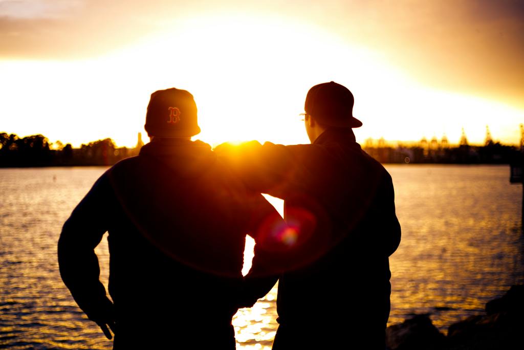 Two friends standing by a lakeside at sunset, with one person placing an arm around the other in a warm gesture of companionship.
